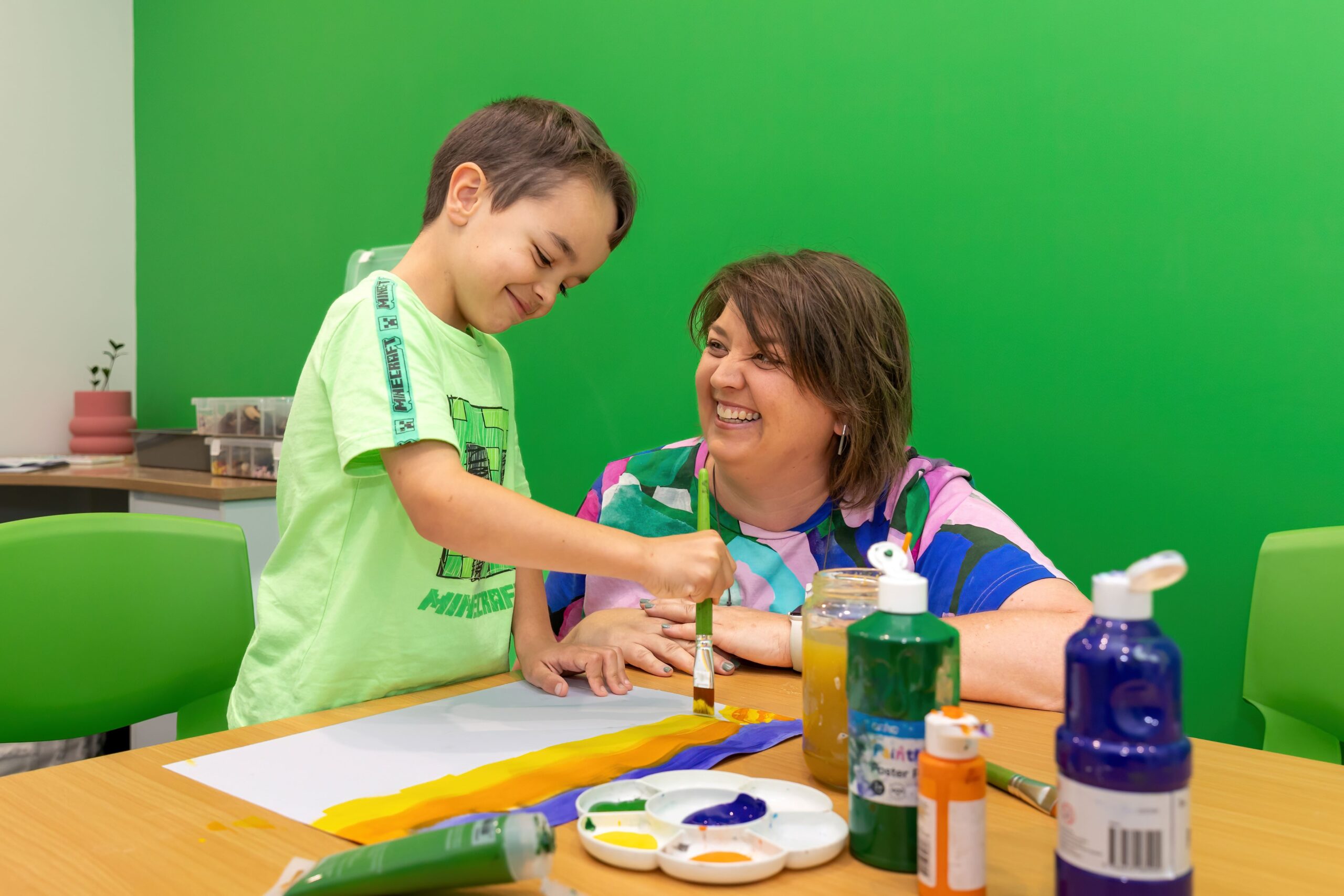 A child wearing a green shirt is painting with our Art Therapist, Deb, who is smiling and kneeling behind the table. There are art materials on the table.