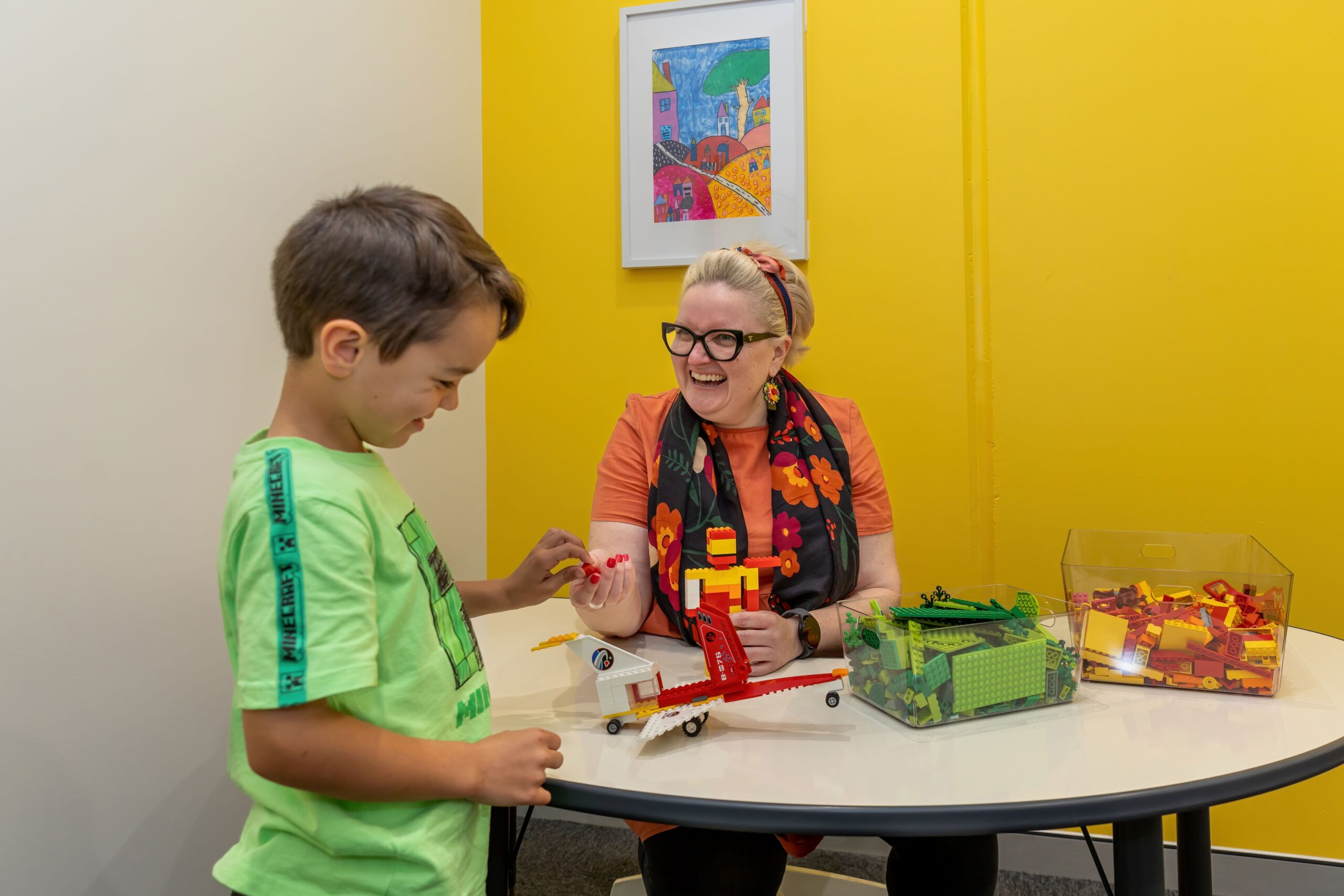 Cicely, the speech therapist, has short blond hair and is wearing glasses. She is wearing an orange shirt with a black scarf. She is playing lego with a young boy, who has brown hair and is wearing a green shirt.