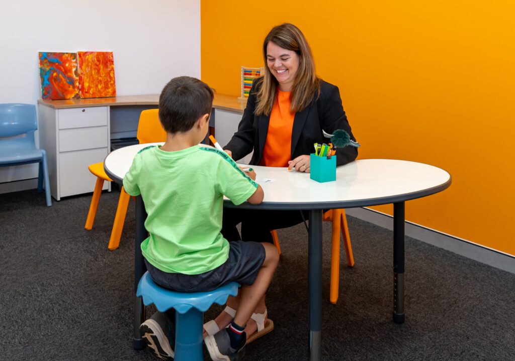 A child is sitting on a wobble stool facing away from the camera and looking at our Speech Therapist, Do who is sitting at a whiteboard table. She is smiling and is wearing an orange shirt and black jacket.