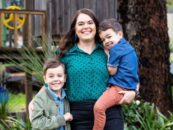 A family of Mum and her two boys. Mum has long brown hair and is smiling. She is wearing a green blouse and black pants. She is holding the younger boy, who has brown hair, is wearing a blue shirt and brick red pants. The other boy has blond hair and is wearing a blue shirt and green jacket. They are all smiling.